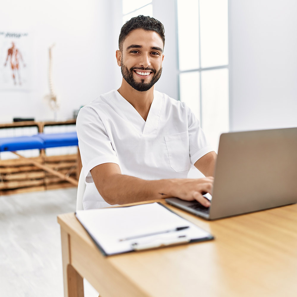 A man sitting at a table with a laptop and smiling at the camera